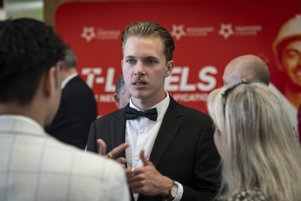 A young man in a black tuxedo and bow tie, with slicked-back hair, stands in conversation, gesturing with his hands. He is in front of a red backdrop with white text and star designs.
