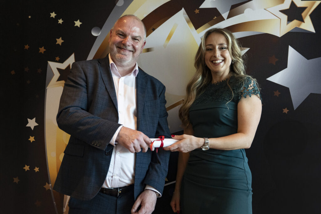 A smiling man in a suit and a smiling woman with long brown hair in a green dress jointly hold a rolled-up certificate. They are against a star-patterned background.