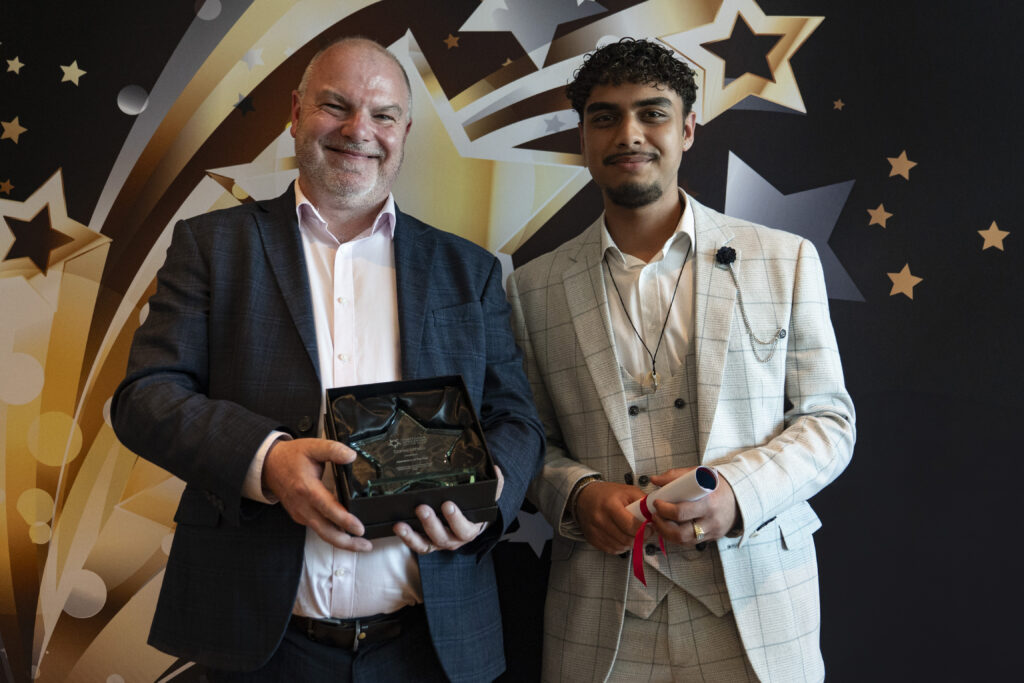 A smiling man in a suit holds a boxed award, standing next to a man in a light suit holding a rolled-up certificate, against a star-patterned background.