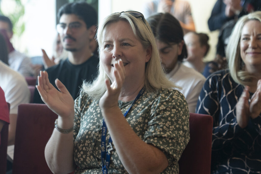 A smiling woman with blonde hair, wearing a floral top and a "STAFF" lanyard, claps with others in an audience.