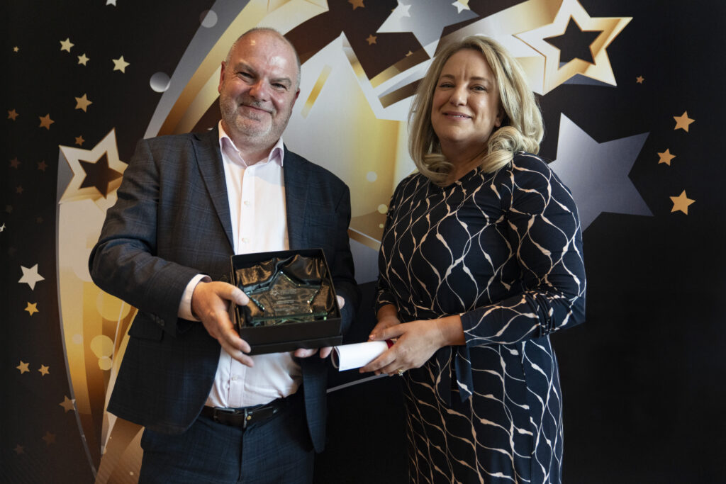 A smiling man in a suit holds a boxed award, standing next to a smiling woman with blonde hair in a patterned dress, who holds a rolled-up certificate. They are against a star-patterned background.