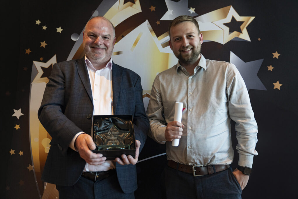  A smiling man in a suit and a smiling man in a light shirt holding a certificate, against a star-patterned background.