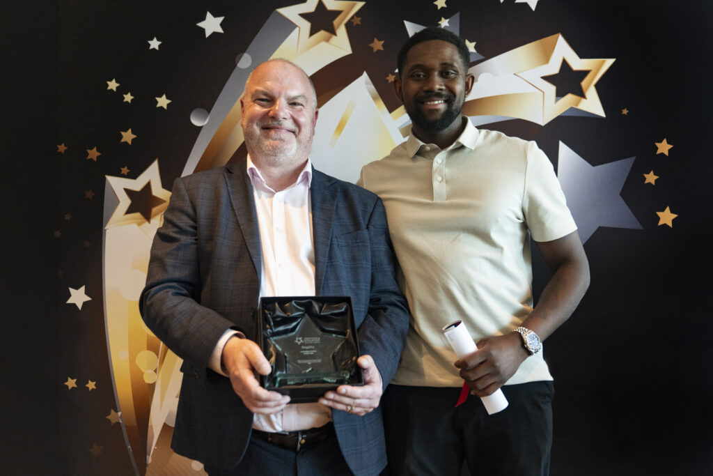 A smiling man in a suit holding an award and a smiling man in a polo shirt holding a certificate, against a star-patterned background.