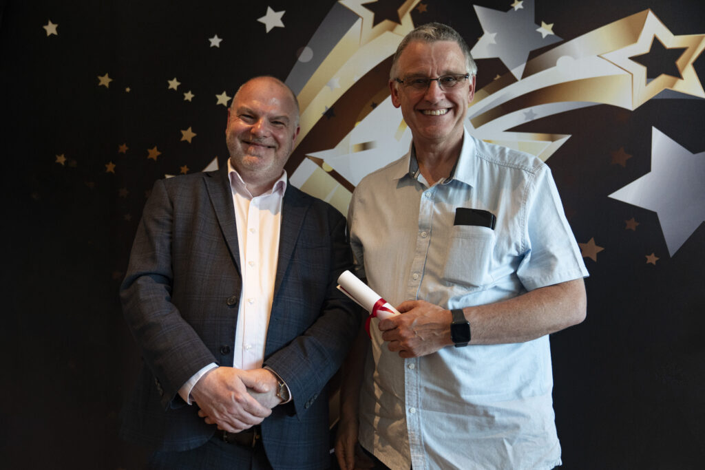  A smiling man in a suit and a smiling man in a light shirt holding a certificate, against a star-patterned background.