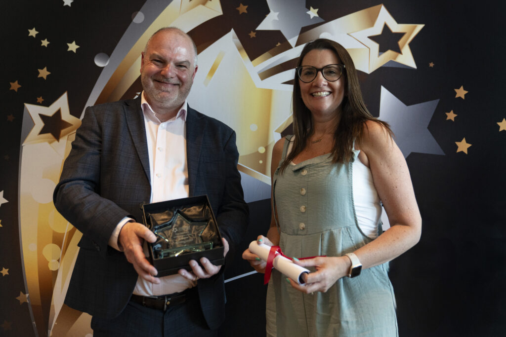A smiling man in a suit holding an award and a smiling woman in overalls holding a certificate, against a star-patterned background.