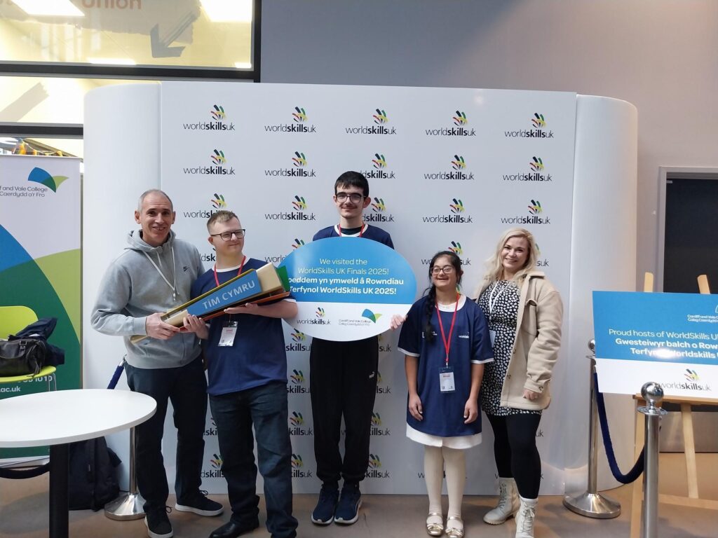A group of five individuals standing in front of a branded backdrop with the WorldSkills UK logo repeated across it. Two people are holding large props: one is a rectangular sign reading “TIM CYMRU,” and another is a circular sign with text in Welsh about WorldSkills UK 2025. There is a small round table on the left and a freestanding banner with colorful graphics. On the right, there is a sign on an easel with more text about WorldSkills UK.