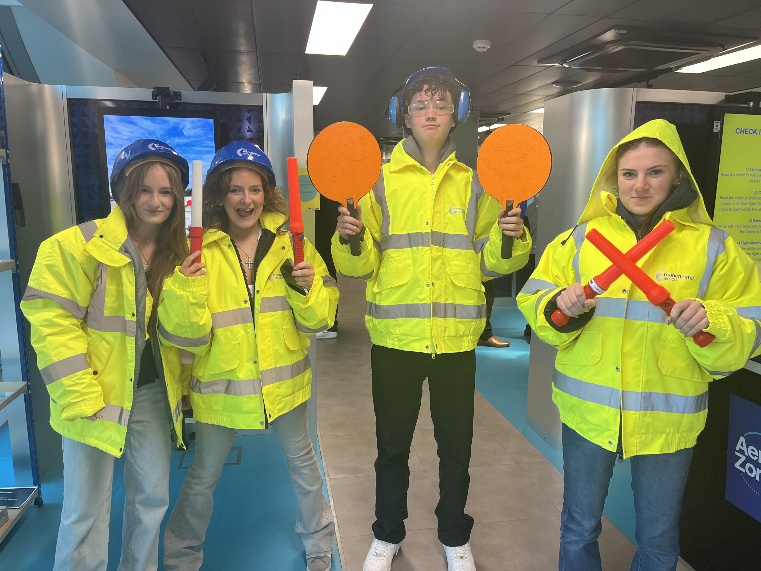 Four individuals wearing yellow high-visibility jackets and safety gear, including helmets and ear defenders, standing indoors. They are holding bright orange and red signalling batons and discs, simulating aircraft marshalling procedures. The background includes signage and equipment related to aviation.
