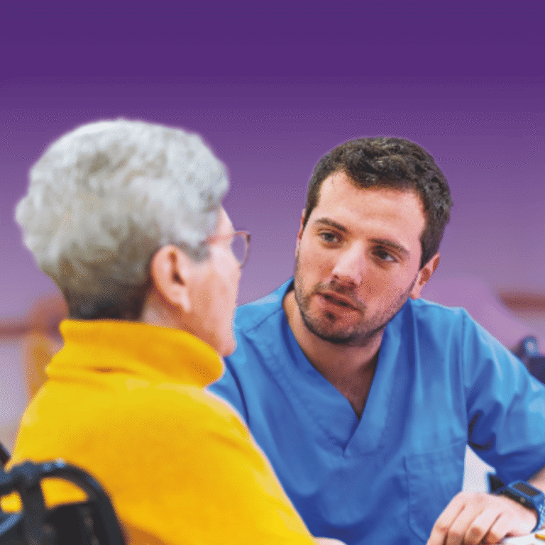 Healthcare professional in scrubs speaking with an older adult in a classroom or training setting.